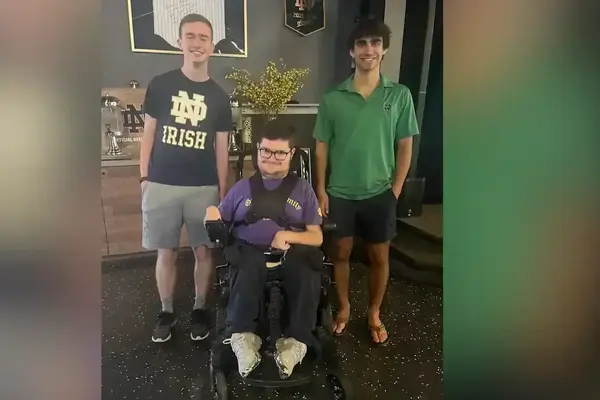 Smiling man in a purple shirt and wheelchair flanked by two standing men. One wears a navy "ND IRISH" shirt, the other a green ND polo.