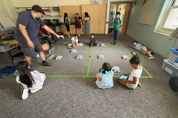 Man in black cap points to a white robot on a green-taped classroom grid. Children sit, watching, some with tablets.