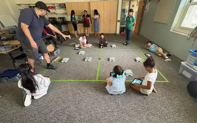 Man in black cap points to a white robot on a green-taped classroom grid. Children sit, watching, some with tablets.